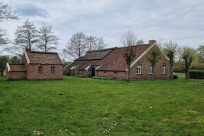 Un bâtiment traditionnel en briques, doté d’un toit à deux versants et d’une petite structure adjacente, le tout entouré d’une zone herbeuse plantée d’arbres.