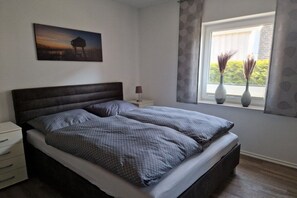 Ein Schlafzimmer mit einem Bett, einem Nachttisch, einer Lampe, einem Fenster mit Vorhängen und einem Bild an der Wand.