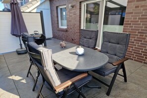 Eine Terrassenanlage mit einem runden Tisch und zwei Stühlen, einem kleinen dekorativen Gegenstand auf dem Tisch und einem Sonnenschirm.