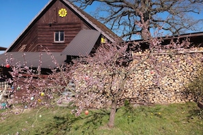 Ein hölzernes Gebäude mit einem geneigten Dach und Schornstein, umgeben von einem Garten mit blühenden Bäumen und einem Holzstapel.