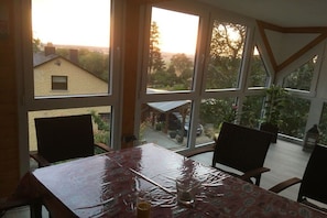 Ein Essbereich mit einem Tisch, der mit einer gemusterten Tischdecke bedeckt ist, großen Fenstern mit Blick nach draußen und Holzstühlen.