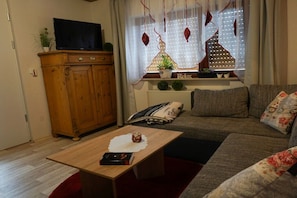 Ein Wohnzimmer mit einem Holzkabinett, einem Fernseher darauf, einer Couch mit Kissen, einem Couchtisch mit einem Buch und einer kleinen Pflanze sowie einem Fenster mit dekorativen Anhängern.