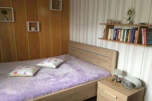 Ein Schlafzimmer mit einem hölzernen Bett, einem Nachttisch mit Lampe und Lautsprecher, einem Bücherregal mit Büchern und einer Wand mit vertikalen Streifen.
