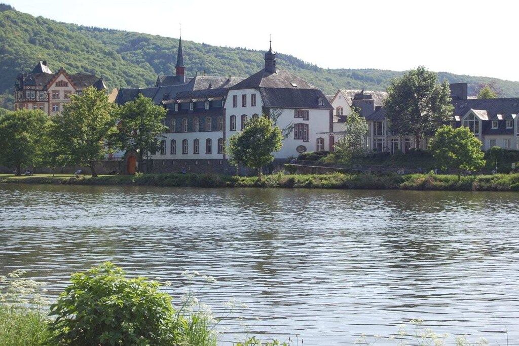 A riverside view with traditional buildings, a church steeple, and a hill in the background.