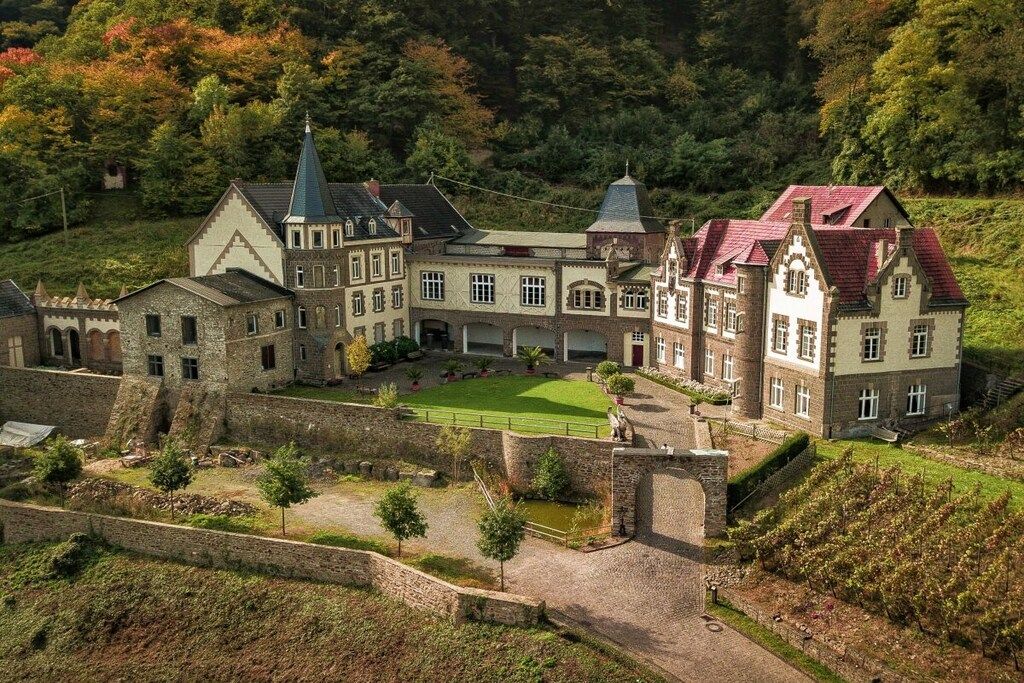 Un grand bâtiment historique doté de plusieurs ailes et d’une cour intérieure, entouré d’un mur de pierre et d’une végétation luxuriante.