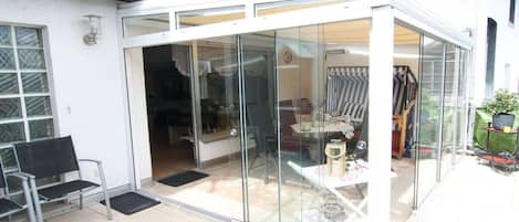 Un espacio de patio con paredes de vidrio, que incluye una mesa, sillas, un banco y una puerta que da a un área interior.