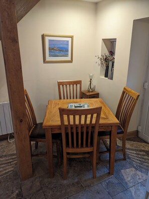 A dining area with a wooden table and chairs, a framed picture on the wall, a small window with flowers, and a door. - Centrally located cosy pet friendly cottage. (Amble)