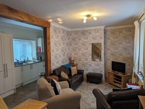 A living room with a sofa, ottoman, and a TV stand. There is a kitchen area visible in the background. - Centrally located cosy pet friendly cottage. (Amble)