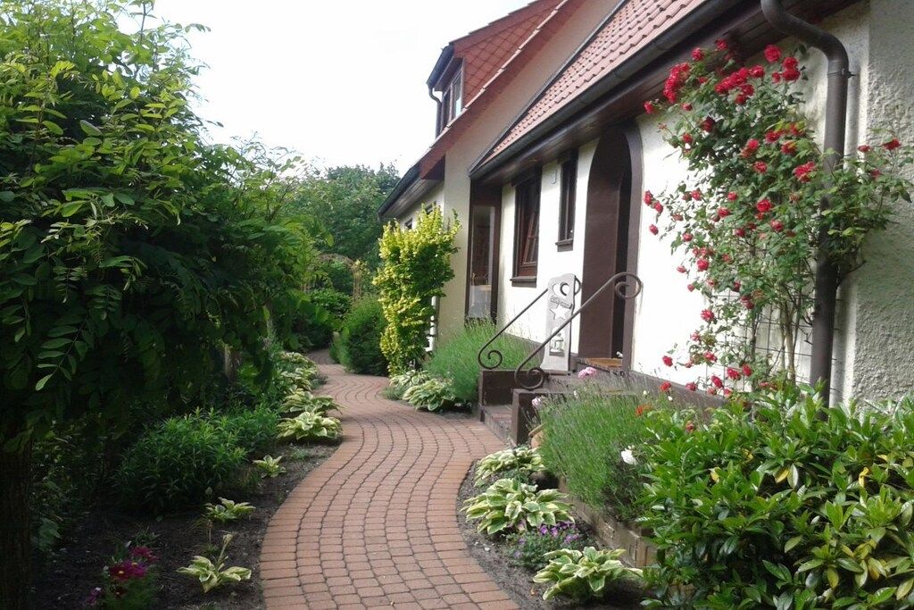 Ein Weg führt zu einem Gebäude mit einem Ziegeldach und einem Garten mit verschiedenen Pflanzen und Blumen.