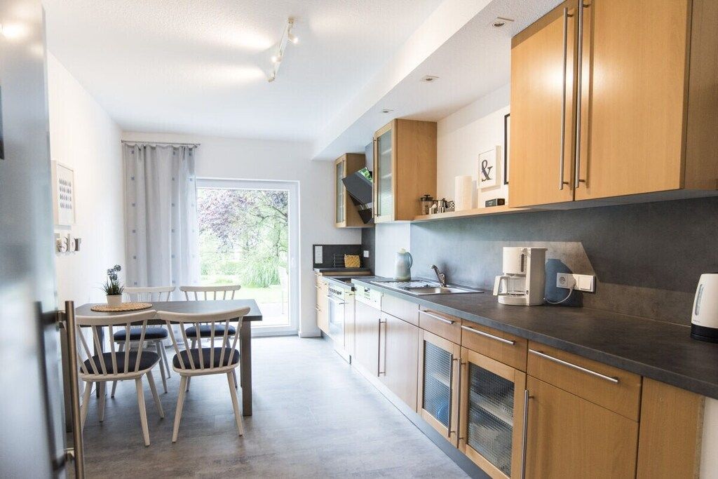 Eine moderne Küche mit Holzausstattung, ein Essbereich mit Tisch und Stühlen sowie ein Blick ins Freie durch ein großes Fenster.