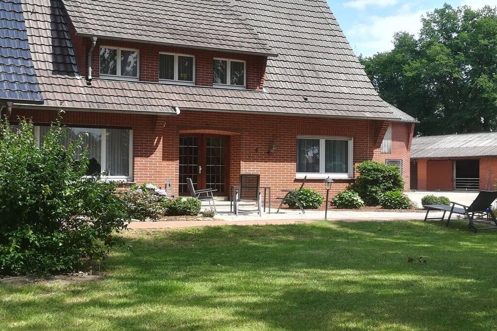 Uma casa de dois andares de tijolo com telhado inclinado, varanda frontal com banco e pequeno gramado.