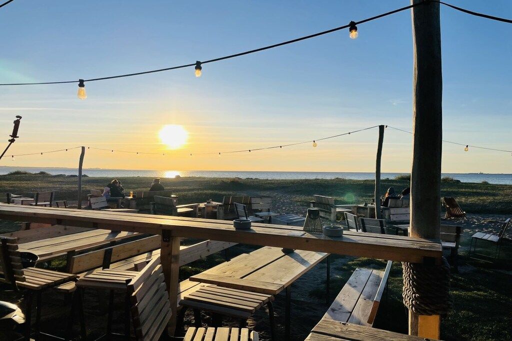Ein Sitzbereich im Freien mit Holzbänken und -tischen, mit Blick auf einen Strand, vor dessen Hintergrund die Sonne untergeht.