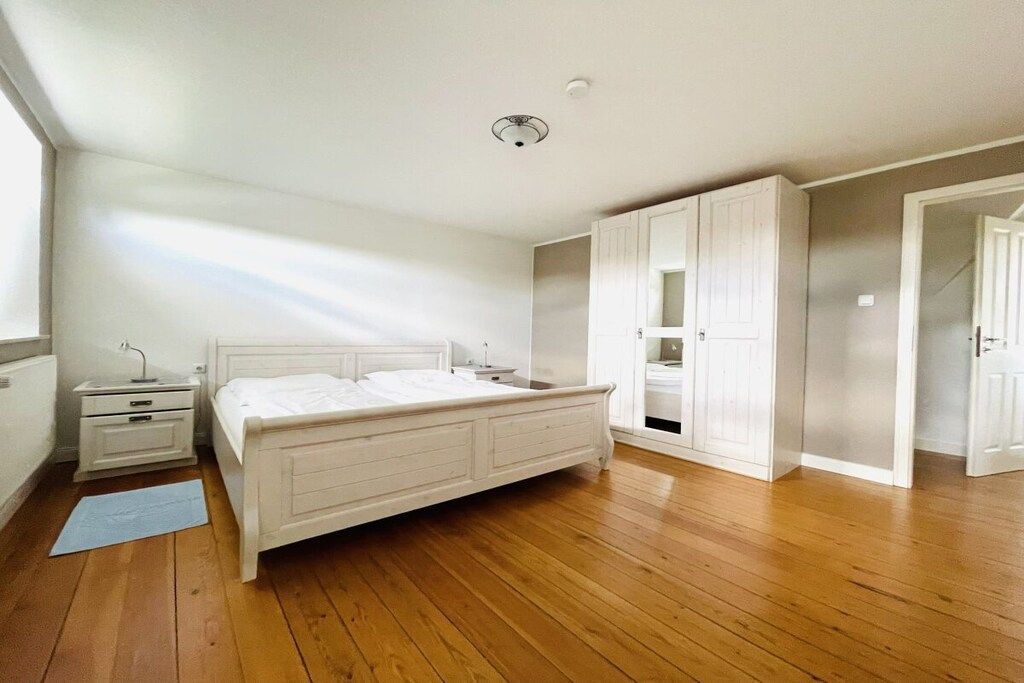 Ein Schlafzimmer mit Holzboden, einem weißen Bett, einem Nachttisch, einem Kleiderschrank und einer Tür zu einem anderen Raum.