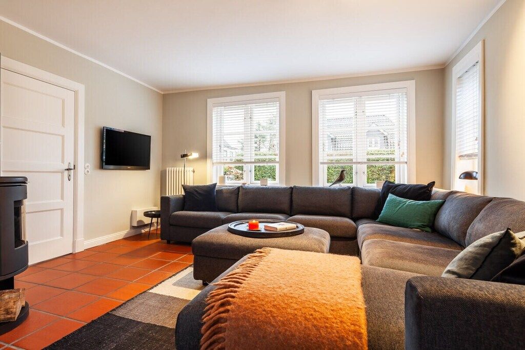 Een moderne woonkamer met een hoekbank, een flatscreen televisie aan de wand en grote ramen met zonwering.