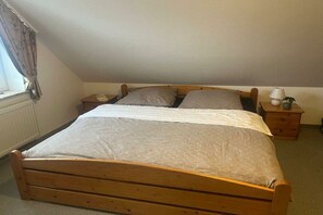 Einzimmer mit einem Bett mit Holzbettgestell, weißer Bettdecke, zwei Kissen, einem Nachttisch mit Lampe und einem Fenster mit Vorhängen.