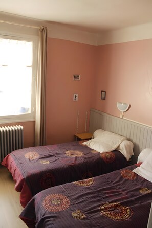 Quarto com duas camas, janela com cortinas, radiador e mesa de cabeceira com abajur.