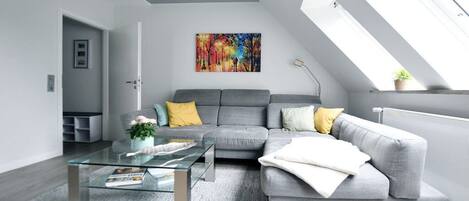 A modern living room with a grey sofa, a glass coffee table, and a colorful painting on the wall.