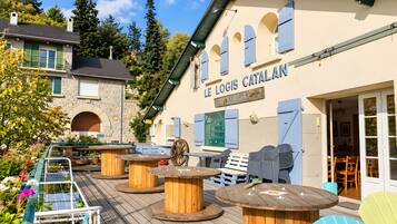 An outdoor seating area with wooden tables and chairs, a stone building with a sign reading 'LE LOGIS CATALAN,' and a view of the surrounding trees and sky.