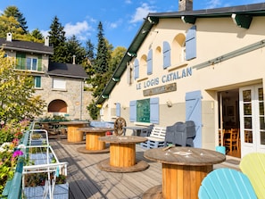 Een terras met houten tafels en stoelen, een stenen gebouw met een bord waarop 'LE LOGIS CATALAN' staat, en een uitzicht op de omliggende bomen en de lucht.