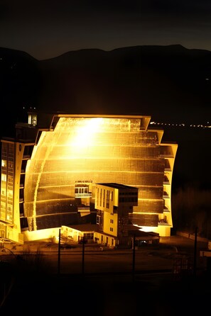 Un grand bâtiment illuminé, doté d’une façade incurvée originale, entouré d’un parking et surplombé par un paysage montagneux.