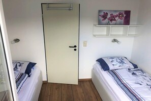 A room with two beds, a door, a shelf with a painting, and a power outlet. - Swallow's Nest Pavilion (Cuxhaven)