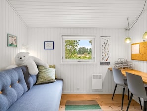 Een gezellige kamer met een blauwe bank, een witte knuffelbeer, een raam met uitzicht op groen, een kleine tafel met stoelen, en een wandlamp.