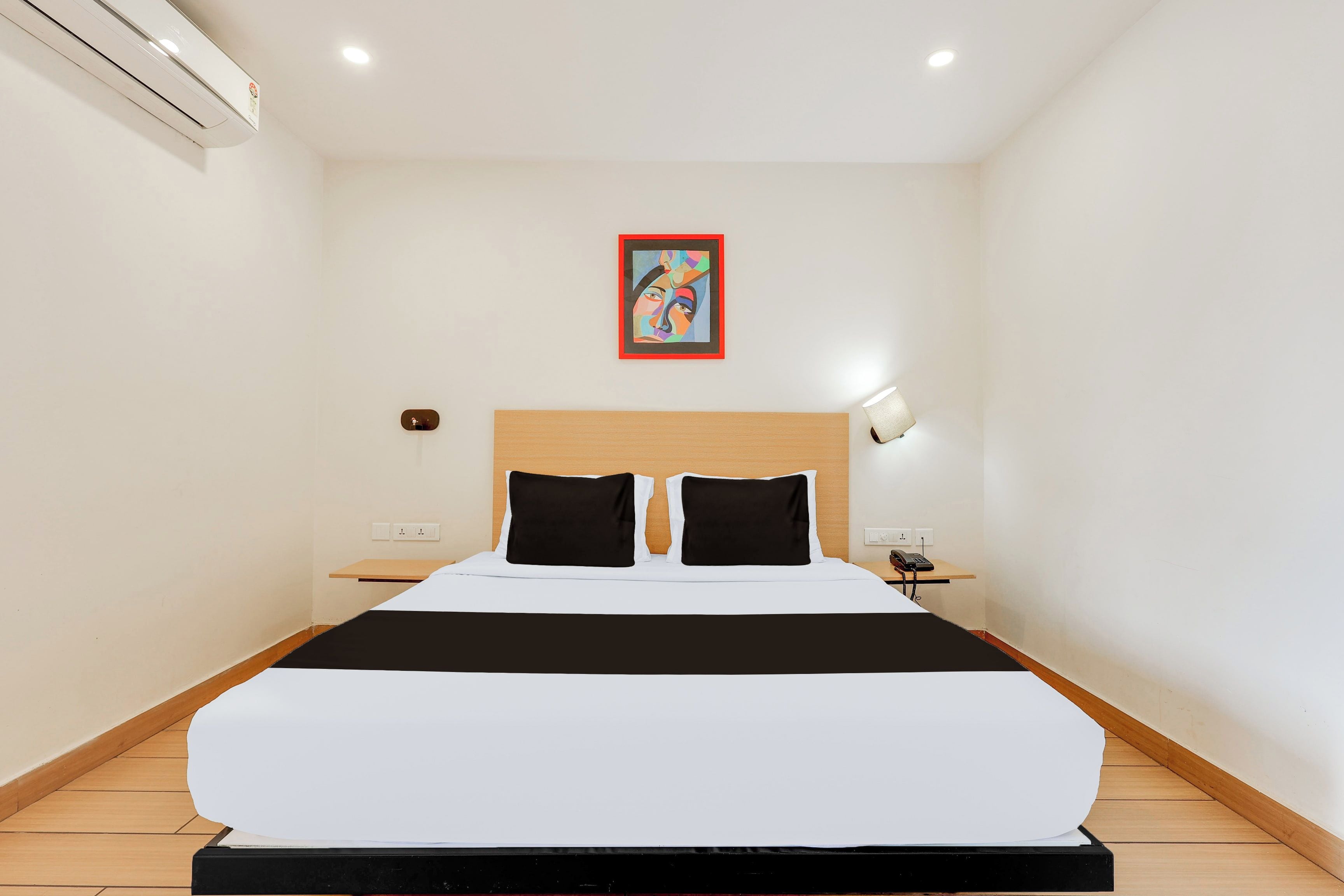 Una habitación de hotel con una cama grande, cabecera, mesita de noche con lámpara y un cuadro colgado en la pared.