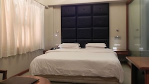 A hotel room with a large bed, a bedside table, a nightstand, a lamp, and a window with curtains.