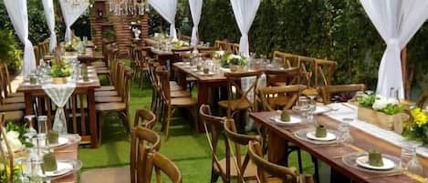 Un área de comedor al aire libre con mesas dispuestas para una comida, rodeada de sillas y cubierta con tela blanca.
