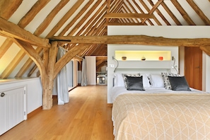 Ein Schlafzimmer mit schrägen Wänden, Holzbalken, einem Bett mit gemusterten Betttextilien und einem Nachttisch mit Lampe.
