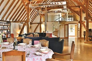 A spacious loft with exposed wooden beams, a dining area with a table set for six, a staircase, and a piano. - The Manor Barn  -  a holiday let that sleeps 10 guests  in 5 bedrooms (Canterbury)