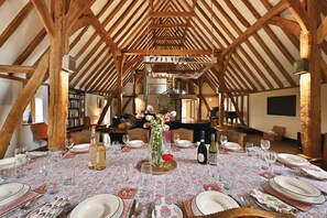 A dining area with a high ceiling and exposed wooden beams, a table set for a meal with plates, glasses, and cutlery, and a vase with flowers in the center. - The Manor Barn  -  a holiday let that sleeps 10 guests  in 5 bedrooms (Canterbury)