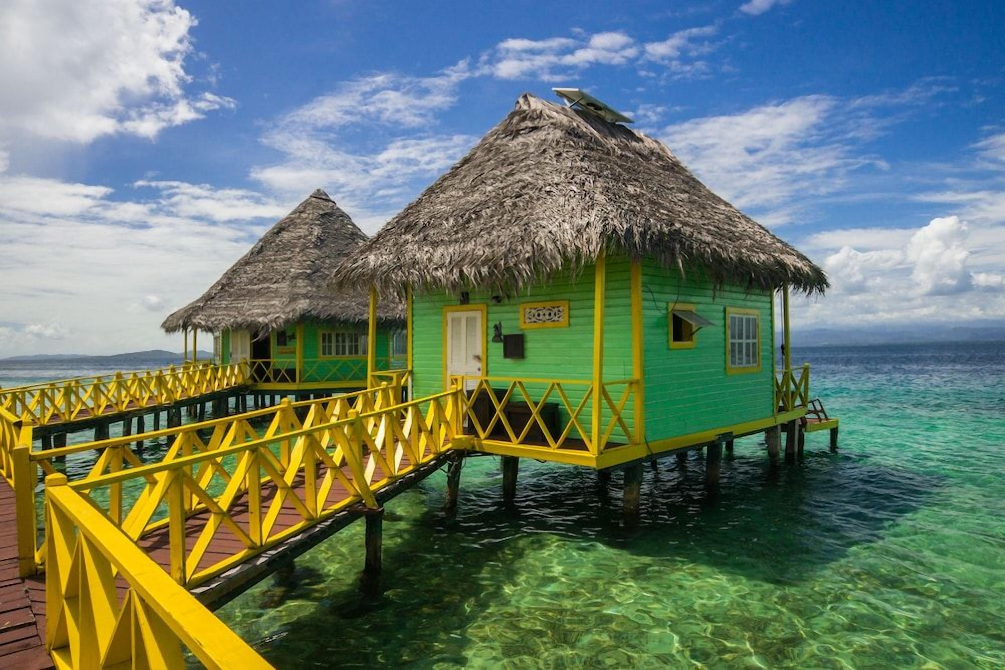 Dos bungalows sobre el agua con techo de paja y barandilla amarilla, unidos por una pasarela de madera, al pie de un cielo azul despejado.