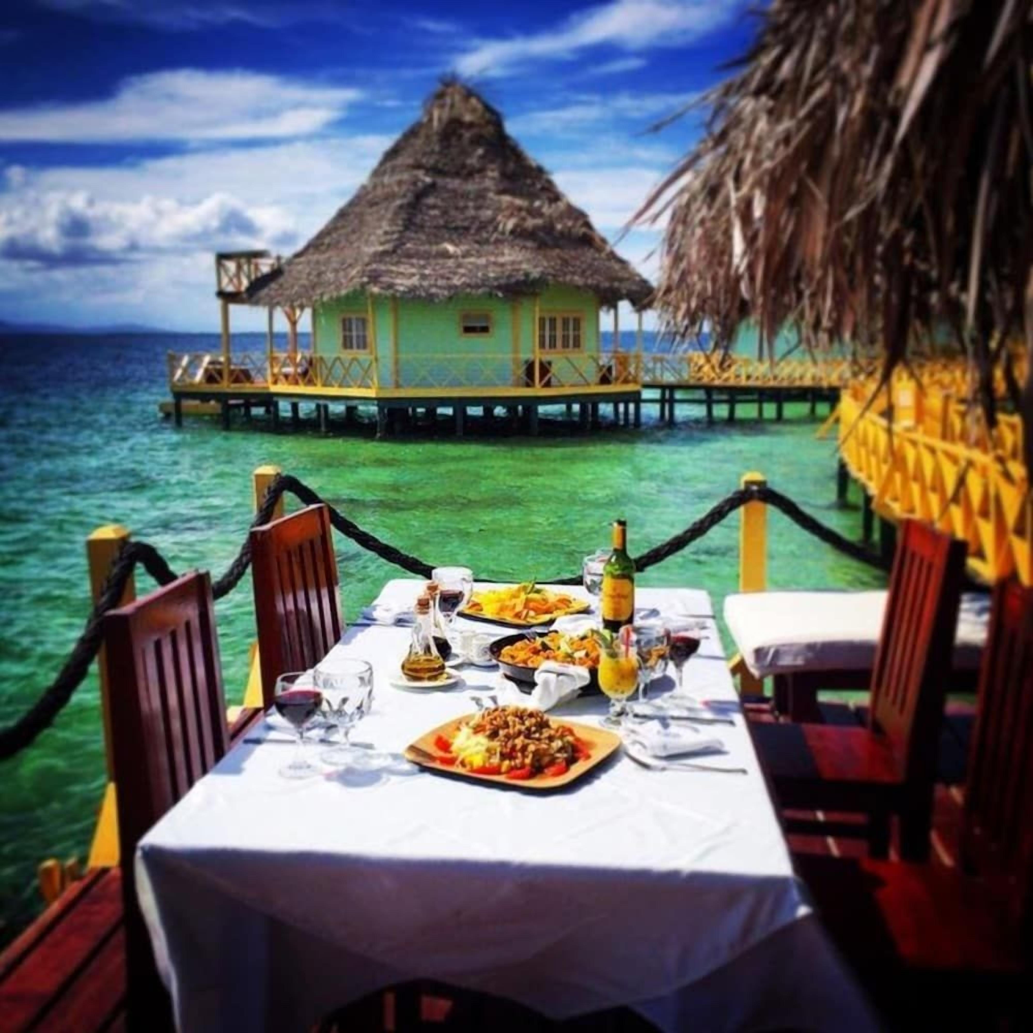 Mesa de comedor dispuesta con comida y bebidas, con vista al mar turquesa y un edificio con techo de paja.