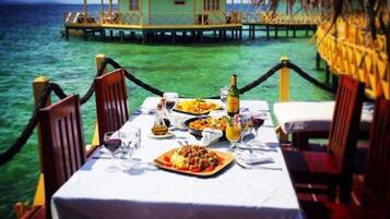 Mesa de comedor dispuesta con comida y bebidas, con vista al mar turquesa y un edificio con techo de paja.