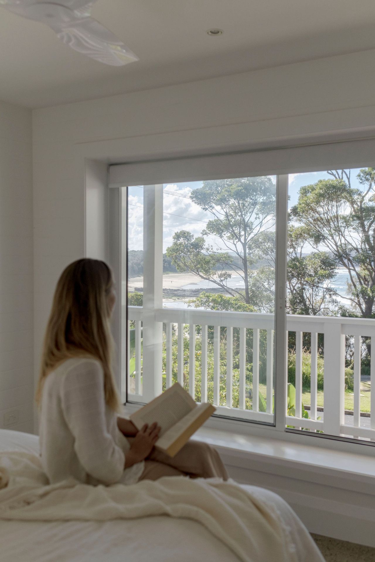 Une personne assise près d’une grande fenêtre, avec vue sur des arbres et une plage.