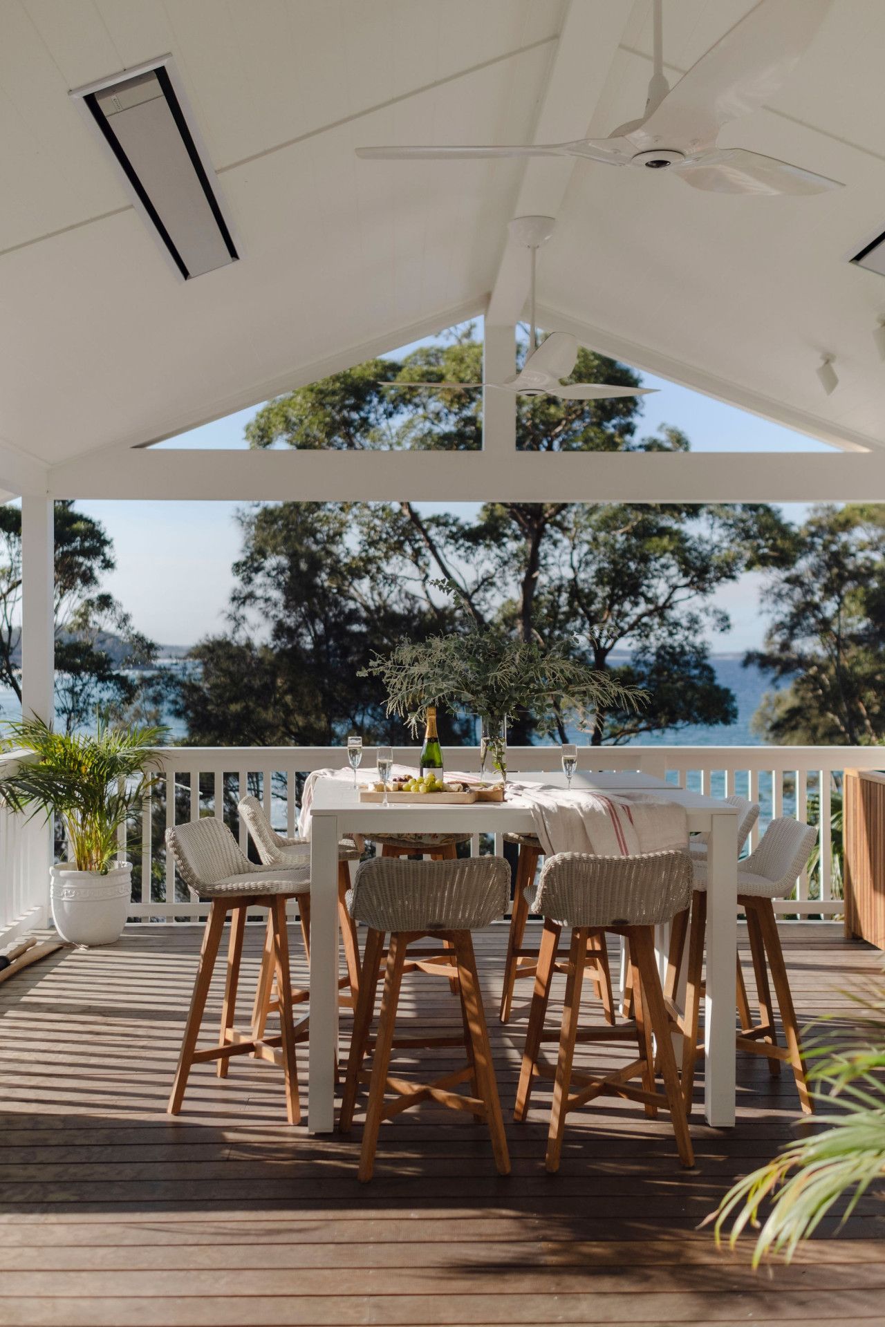 An outdoor dining area with a white table and wicker chairs, overlooking a body of water and trees.