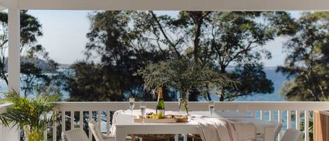 An outdoor dining area with a white table and wicker chairs, overlooking a body of water and trees.