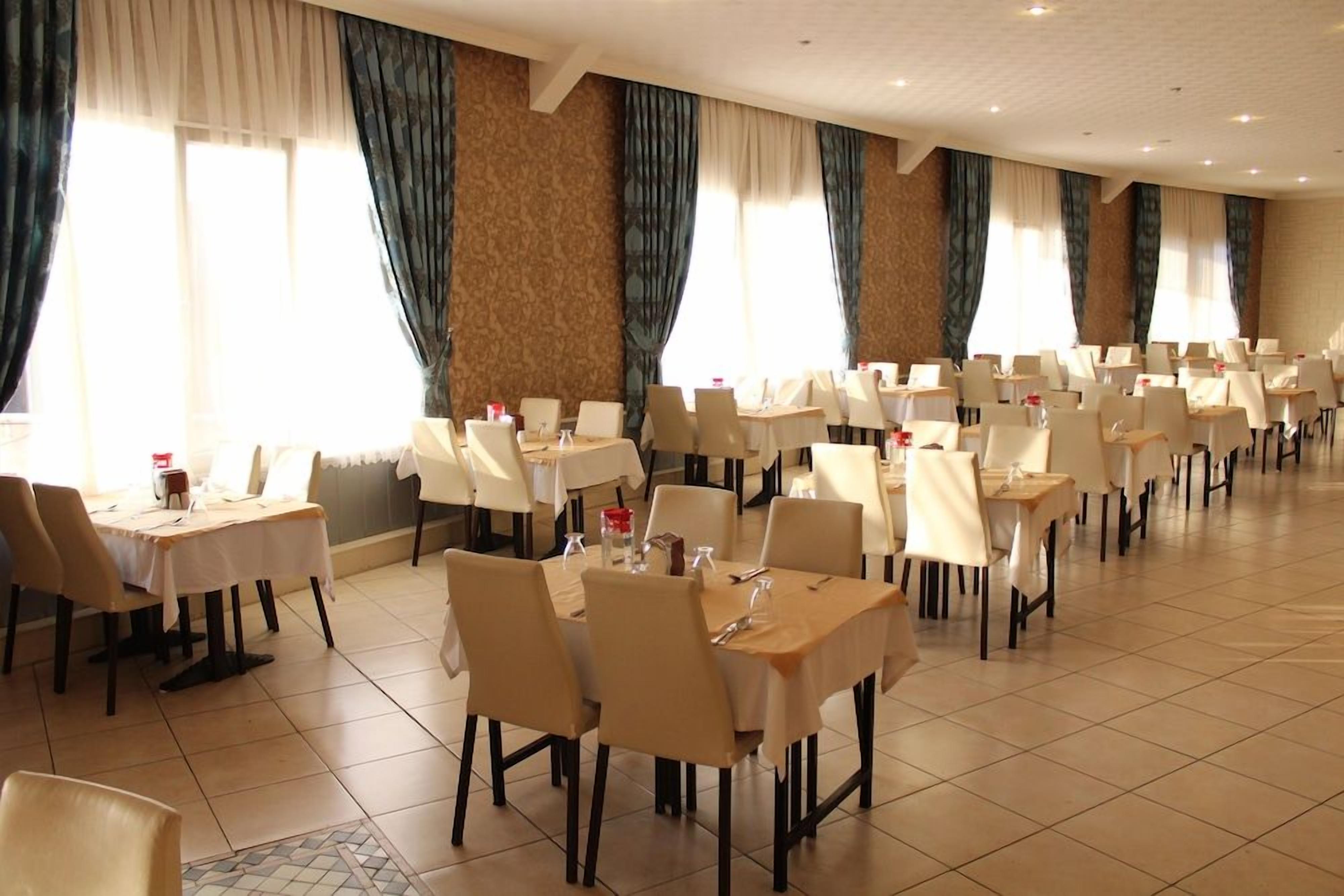Ein Hotel-Speisesaal mit mehreren Tischen und Stühlen, großen Fenstern mit Vorhängen und einem Fliesenboden.