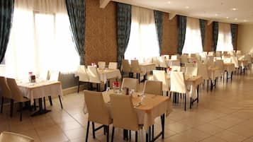 Ein Hotel-Speisesaal mit mehreren Tischen und Stühlen, großen Fenstern mit Vorhängen und einem Fliesenboden.