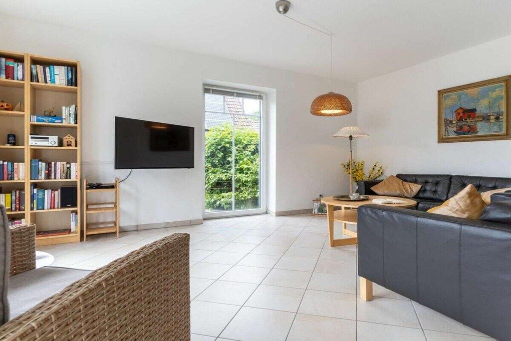 En moderne stue med sofa, fjernsyn, bogreol og udsigt til grønne områder.