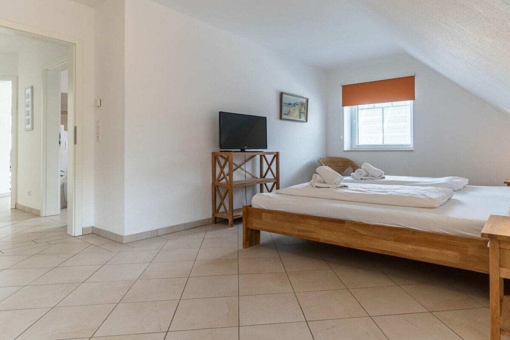 A bedroom with a wooden bed, a television on a wooden stand, a window with blinds, and a door leading to another room.