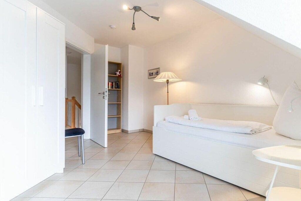 Quarto moderno e bem iluminado, com sofá branco, uma mesinha e um abajur. Há uma estante e uma porta que dá para outro cômodo.
