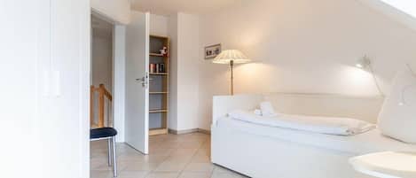 Quarto moderno e bem iluminado, com sofá branco, uma mesinha e um abajur. Há uma estante e uma porta que dá para outro cômodo.