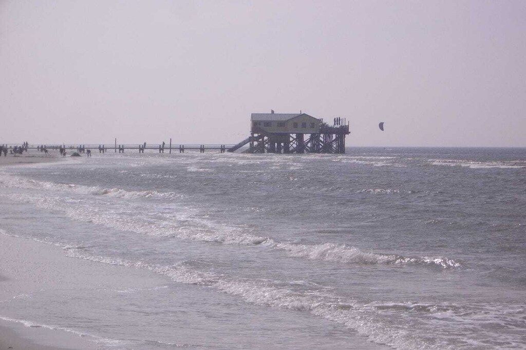A beach with a pier extending into the water, a building on the pier, and a person kite surfing in the distance.