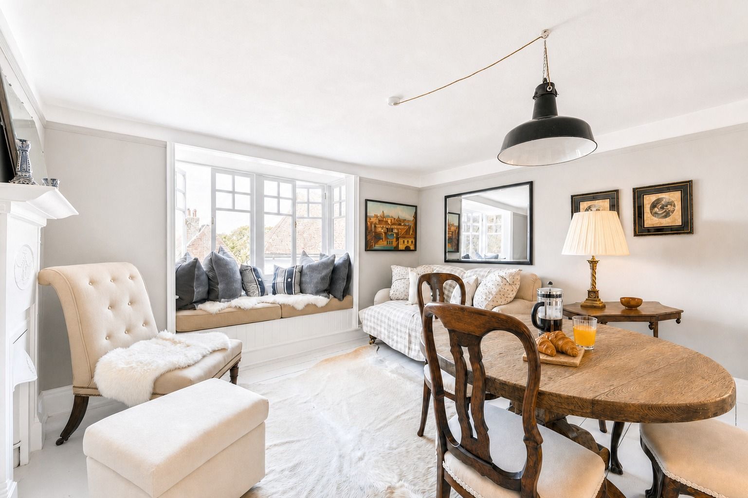 A cozy room with a large window, a dining table with chairs, a sofa, and a chair with a fur throw.