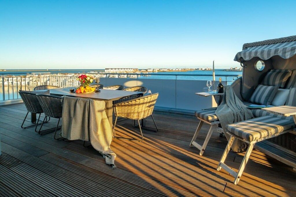 A deck with a table and chairs, a sofa, and a view of the sea and buildings.
