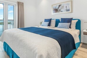 Ein Hotelzimmer mit einem großen Bett, Meerblick durch eine Schiebetür und einem gerahmten Bild an der Wand.