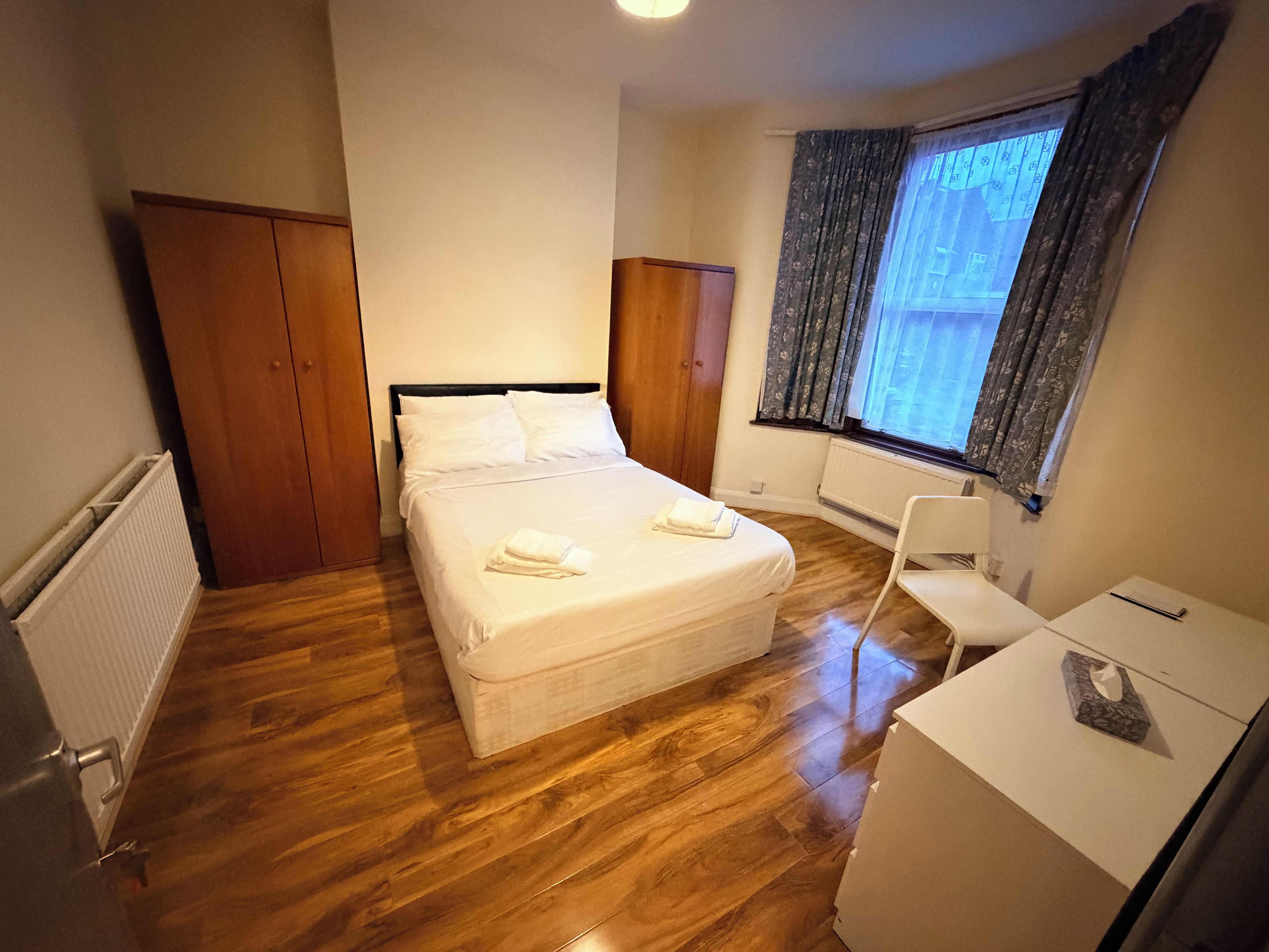Une chambre d’hôtel avec un lit, un bureau, une chaise, une armoire et une fenêtre avec des rideaux.