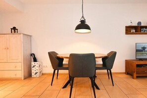 A dining area with a round table, four chairs, a hanging light fixture, a cabinet, a small cabinet with a decorative item, and a television on a wooden stand. - Holiday apartment in Büsum with swimming pool (Büsum)